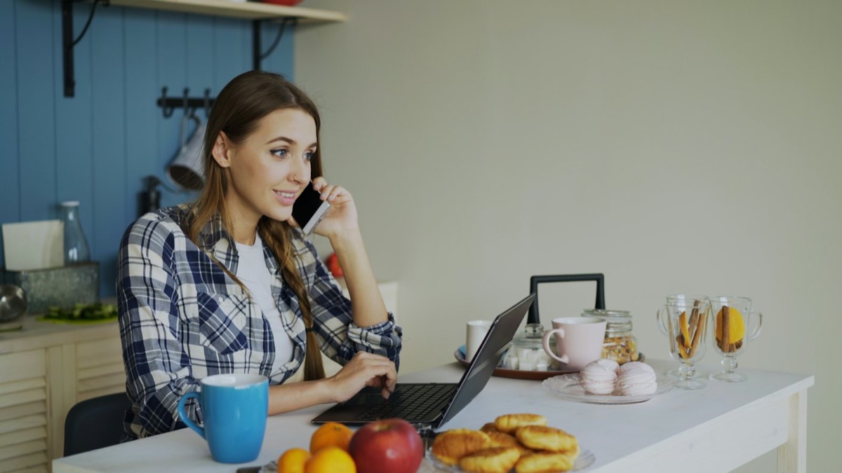 Mulher sorrindo ao telefone enquanto usa o notebook na cozinha